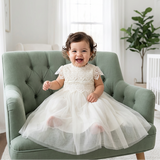 Child in a white dress sitting on a green chair in a bright room with a plant in the background