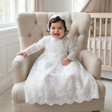 Child in a white lace dress sitting on a beige armchair in a bright room.