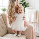 Woman holding a baby in a white dress in a bright room with a crib and plants.
