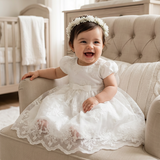 Baby girl in a white lace dress sitting on a beige couch in a nursery.