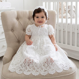 Baby in a white lace dress sitting on a beige chair in a nursery.