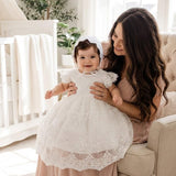 Woman holding a baby in a white lace dress in a bright room with a crib and plants.